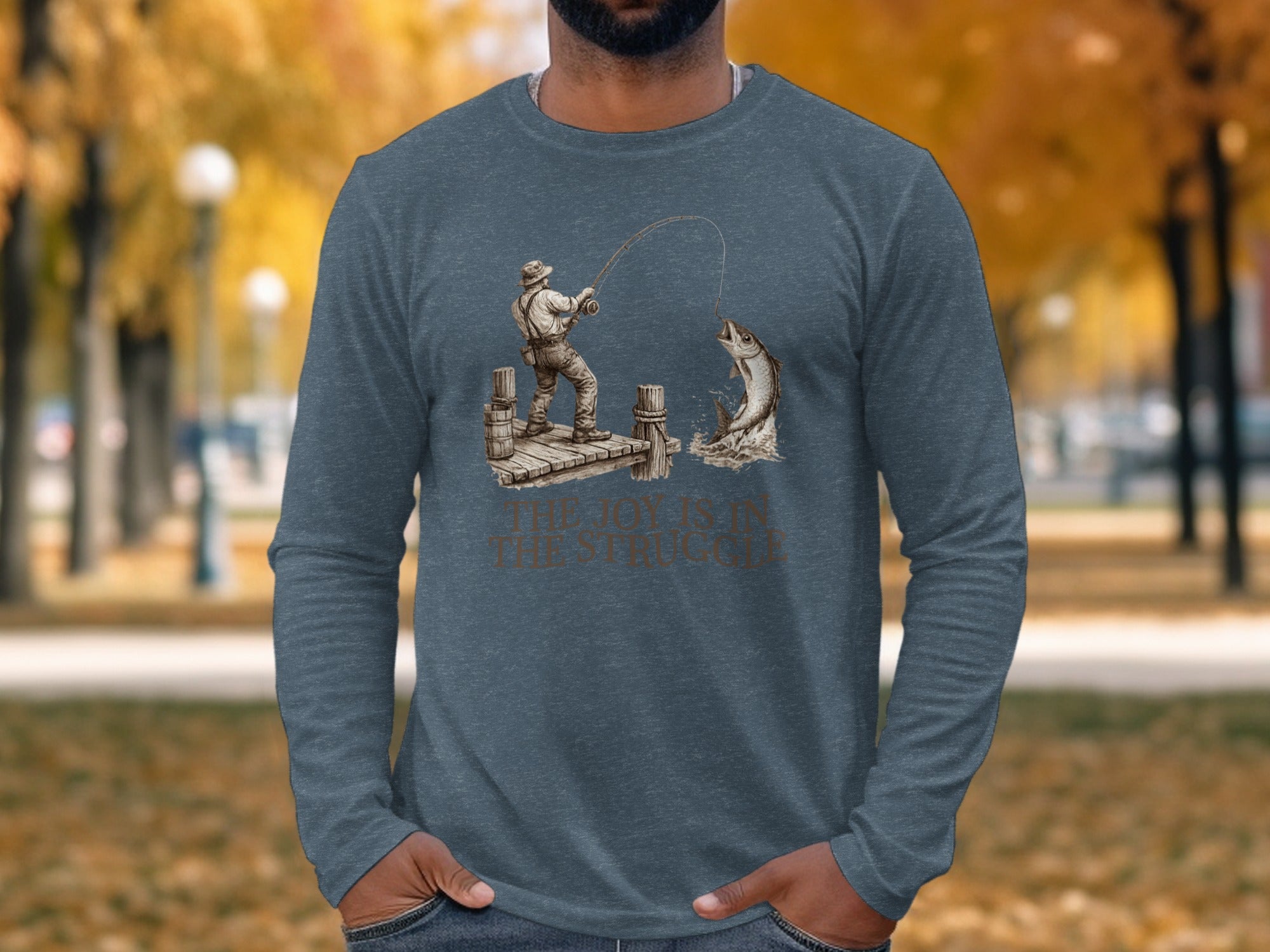 Long sleeve t-shirt featuring a fisherman reeling in a large fish, with text "THE JOY IS IN THE STRUGGLE".