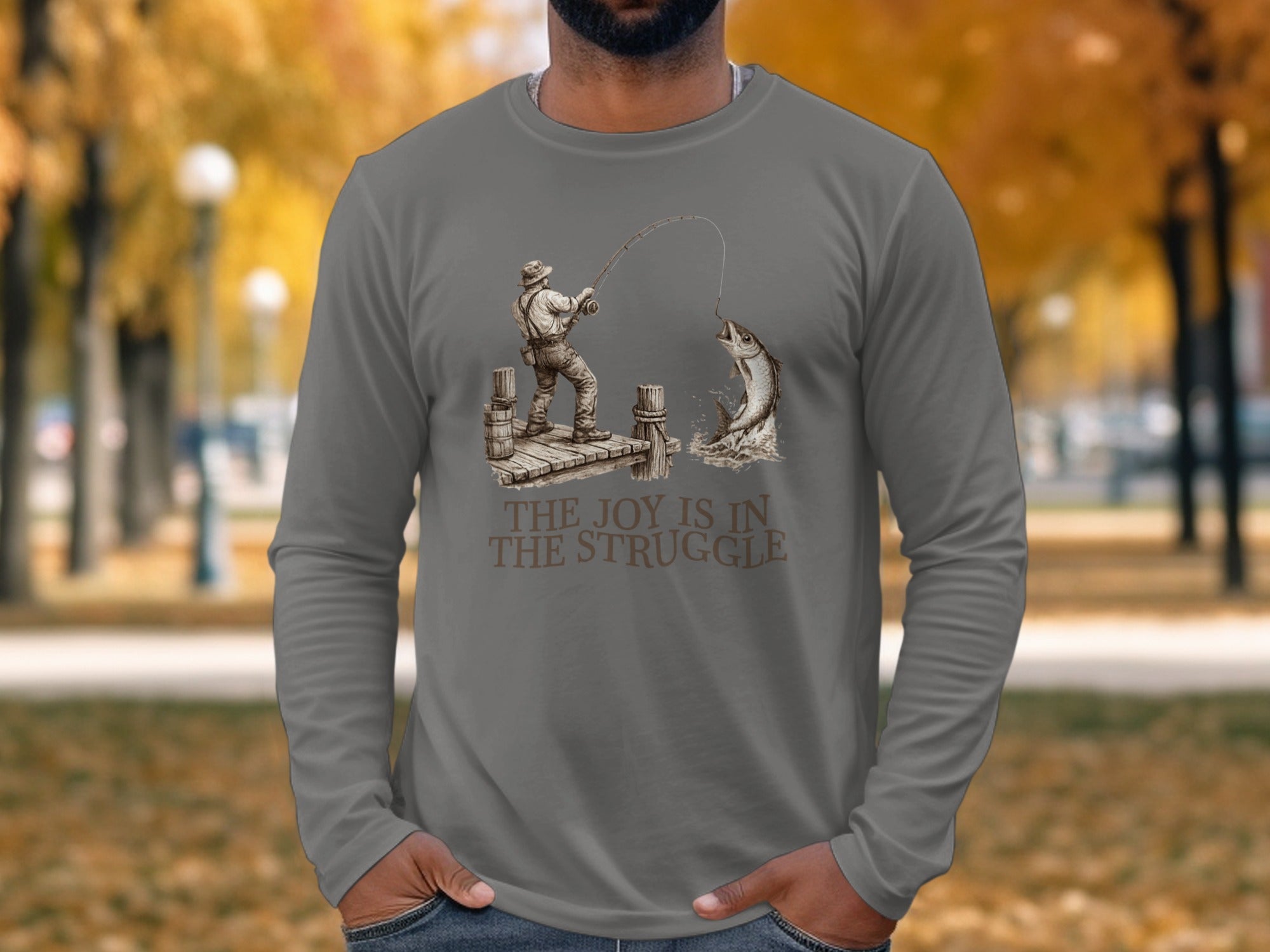 Man in long-sleeve shirt showing a fishing scene with the text "THE JOY IS IN THE STRUGGLE".