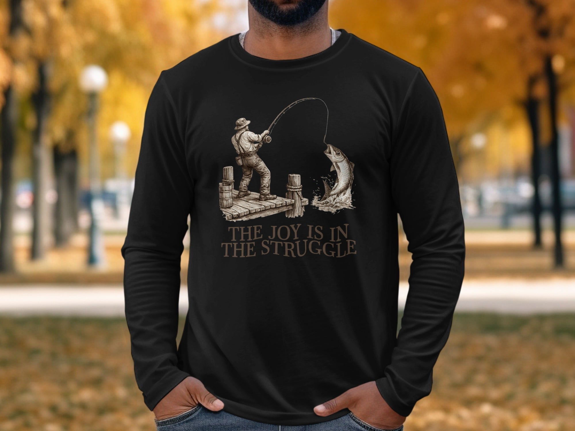 Black long-sleeve shirt with a fisherman catching a large fish, text reads "The Joy is in the Struggle."
